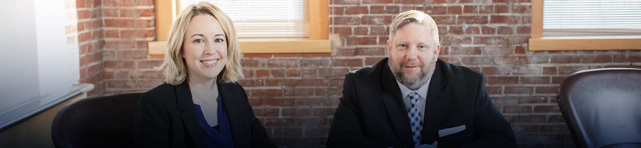 A photograph capturing the interior of the offices, featuring Douglas Ponder and Jaclyn Zimmermann seated and leaning against a glass table. In the background, a brick decorated wall and a window are visible.